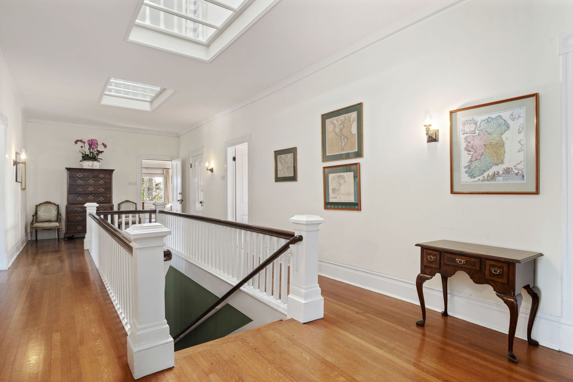 Bright hallway with hardwood floors, skylights, framed art, and a staircase railing.