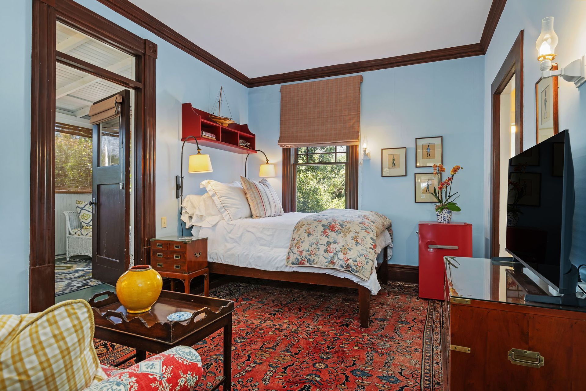 Cozy bedroom with red patterned rug, white bed, wooden furniture, and blue walls with windows.