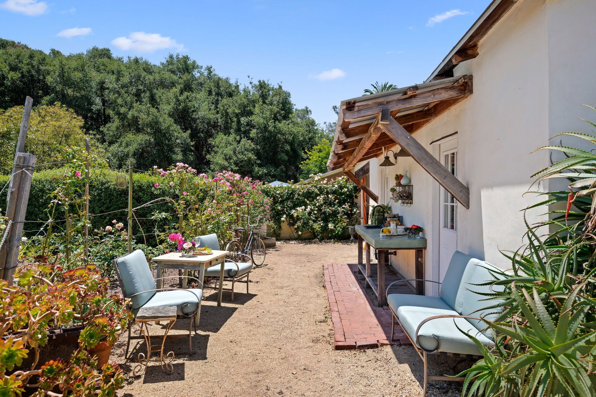 Sunny garden patio beside a white house with table, chairs, potted plants, and lush greenery