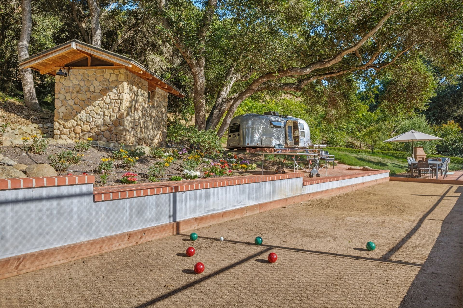 Outdoor pétanque court with scattered balls beside a stone terrace and trees.