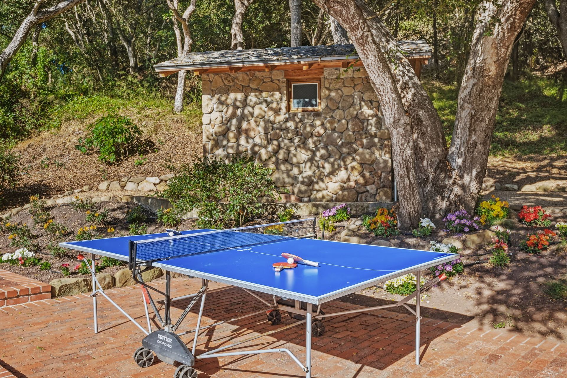 Blue ping-pong table outdoors on a brick patio near a stone wall and trees, with paddles and balls on top.