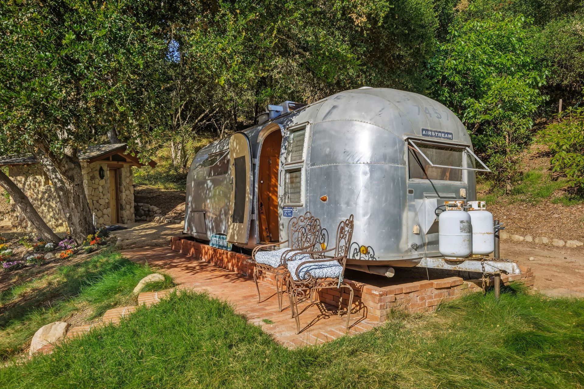 Silver tiny house trailer in a wooded clearing, with propane tanks and outdoor seating.