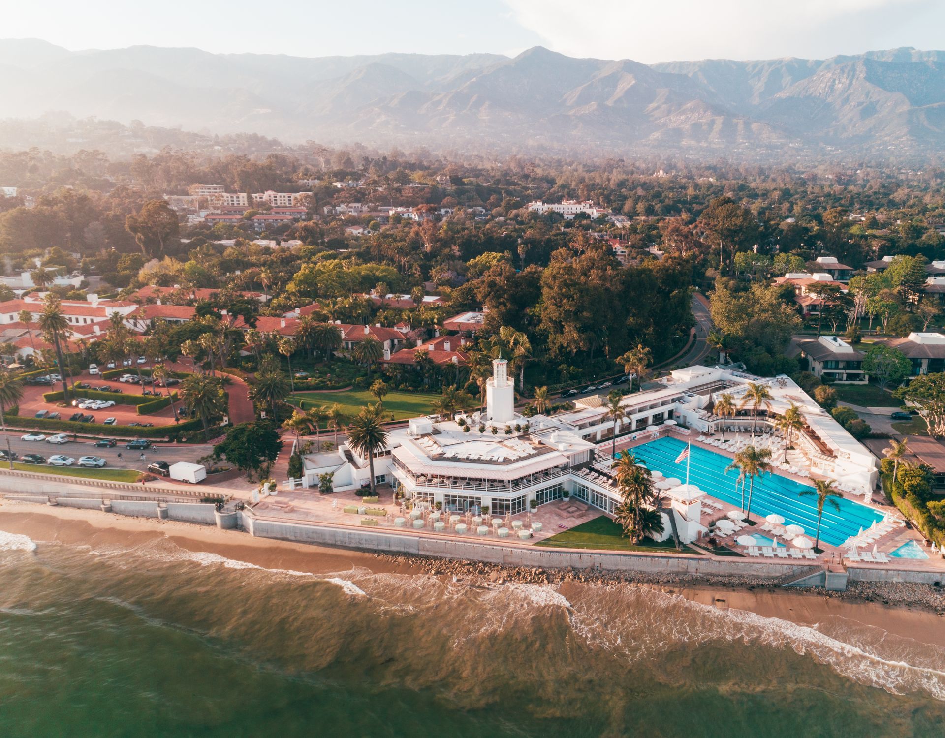 Aerial view of a beach resort with a pool, shoreline, and distant mountains at sunset