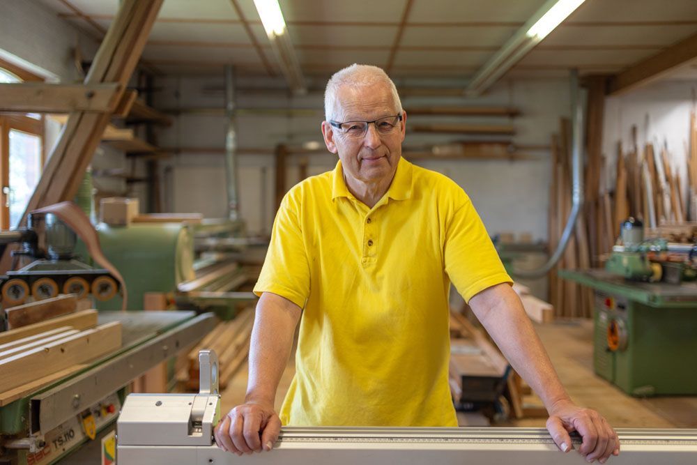 Ein Mann in einem gelben Hemd steht neben einer Maschine in einer Holzwerkstatt.