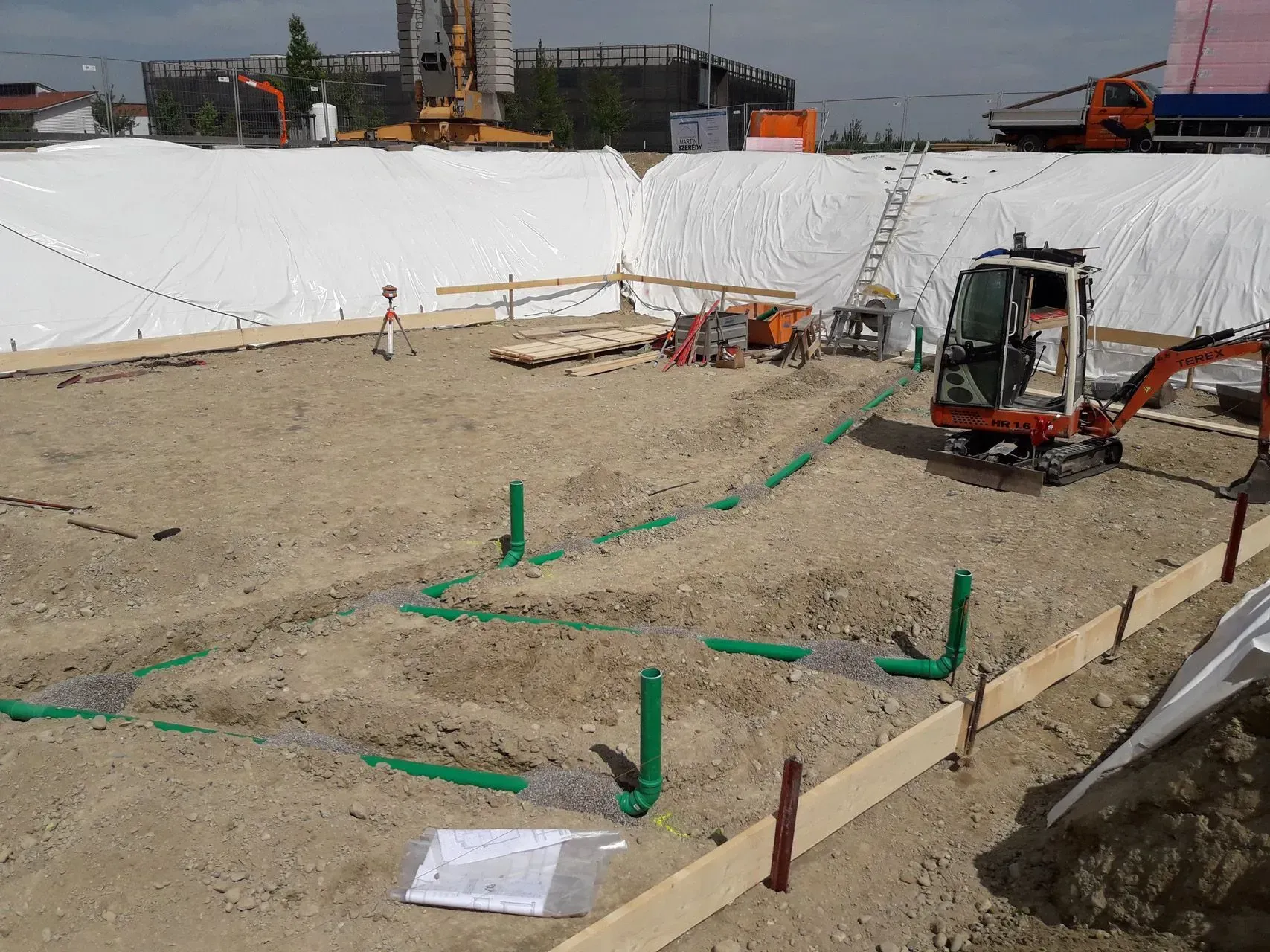 Baustelle mit Bagger auf sandigem Boden, grünen Markierungen und im Hintergrund weißen, mit Planen abgedeckten Böschungen.