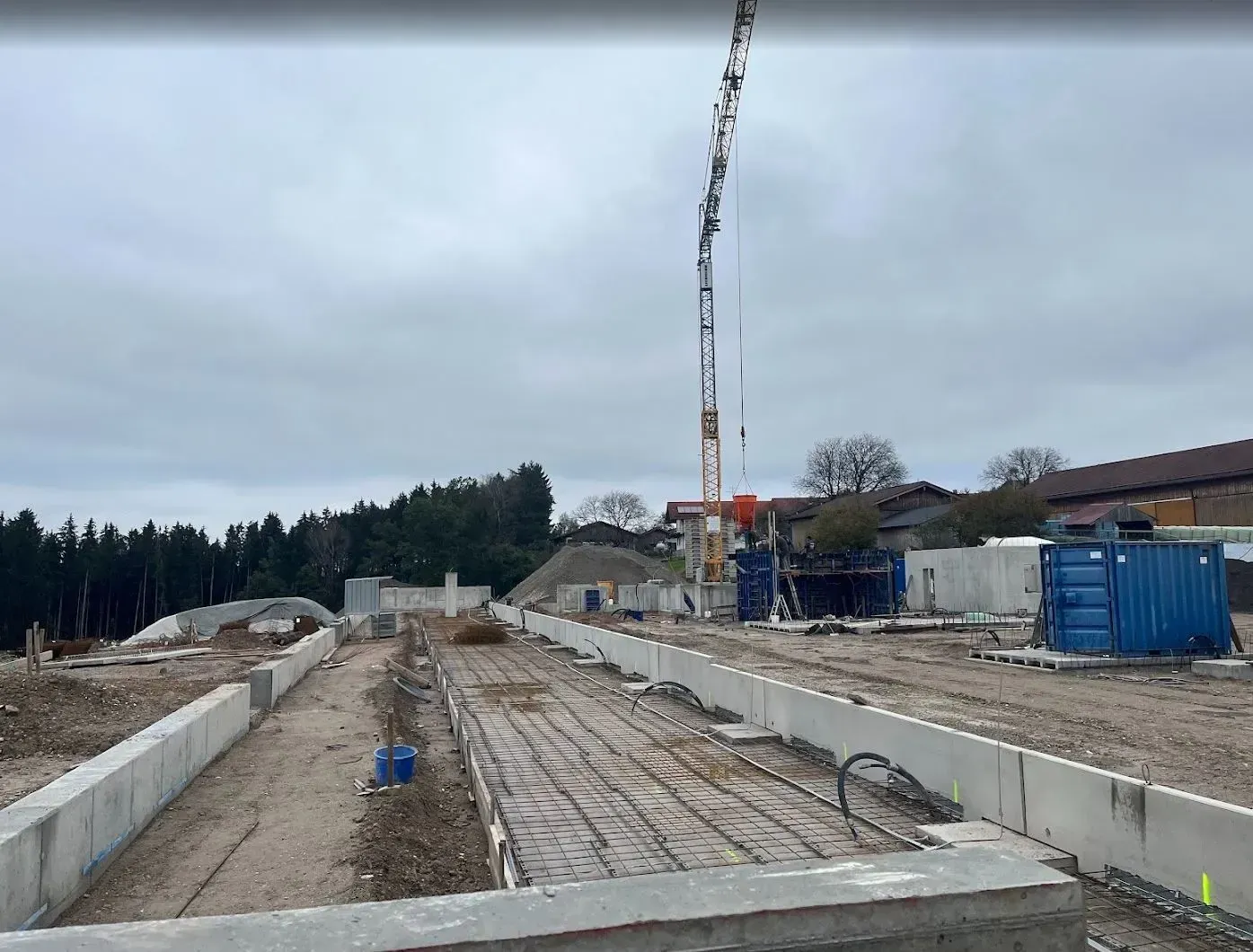 Baustelle mit Betonfundamenten, Kran und blauen Baustellencontainern unter einem bedeckten Himmel