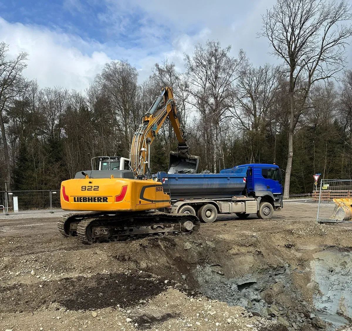 Ein gelber Bagger belädt einen blauen Muldenkipper auf einer schlammigen Baustelle unter bewölktem Himmel.