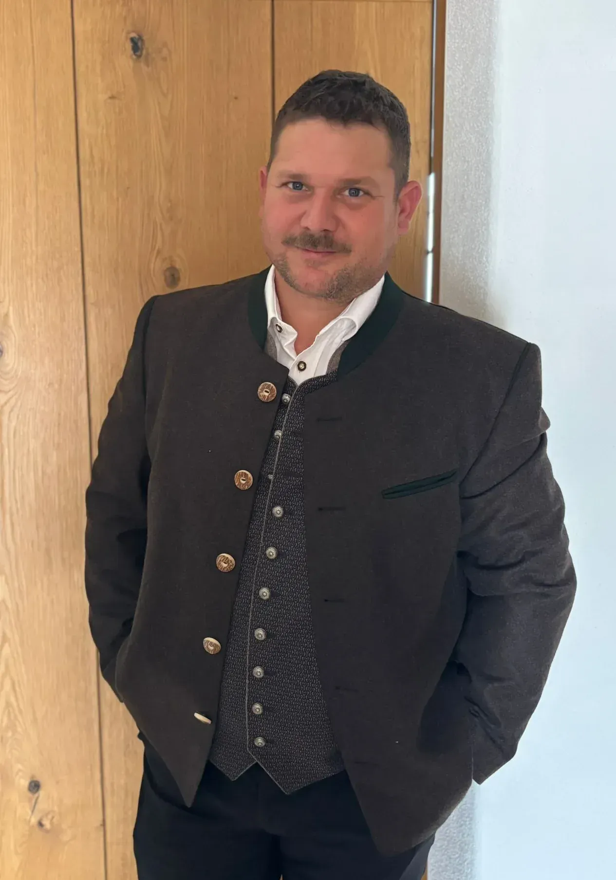 Ein Mann in dunkler Jacke und gemusterter Weste steht in einem Innenraum, lehnt mit einer Hand in der Tasche an einer Tür.