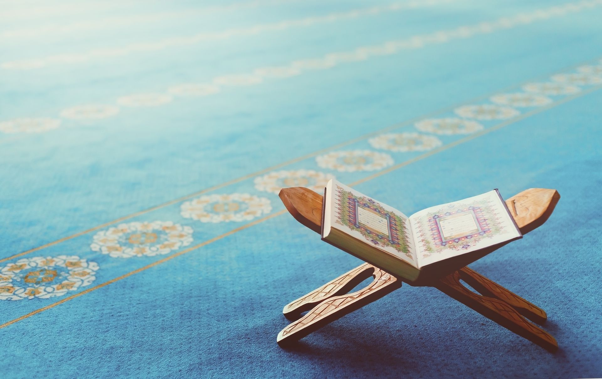 Coran ouvert sur un support en bois, placé sur un tapis de prière bleu à motifs.