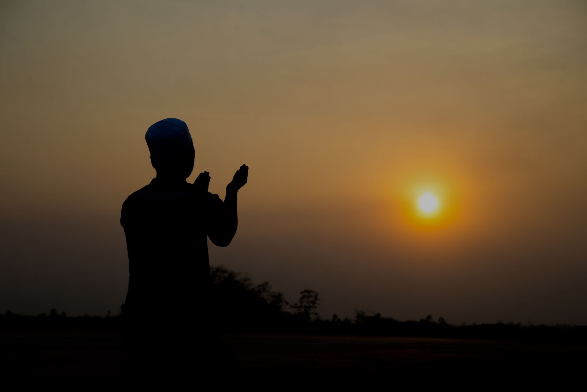 Silhouette d'un homme en prière face à un coucher de soleil, portant une casquette.