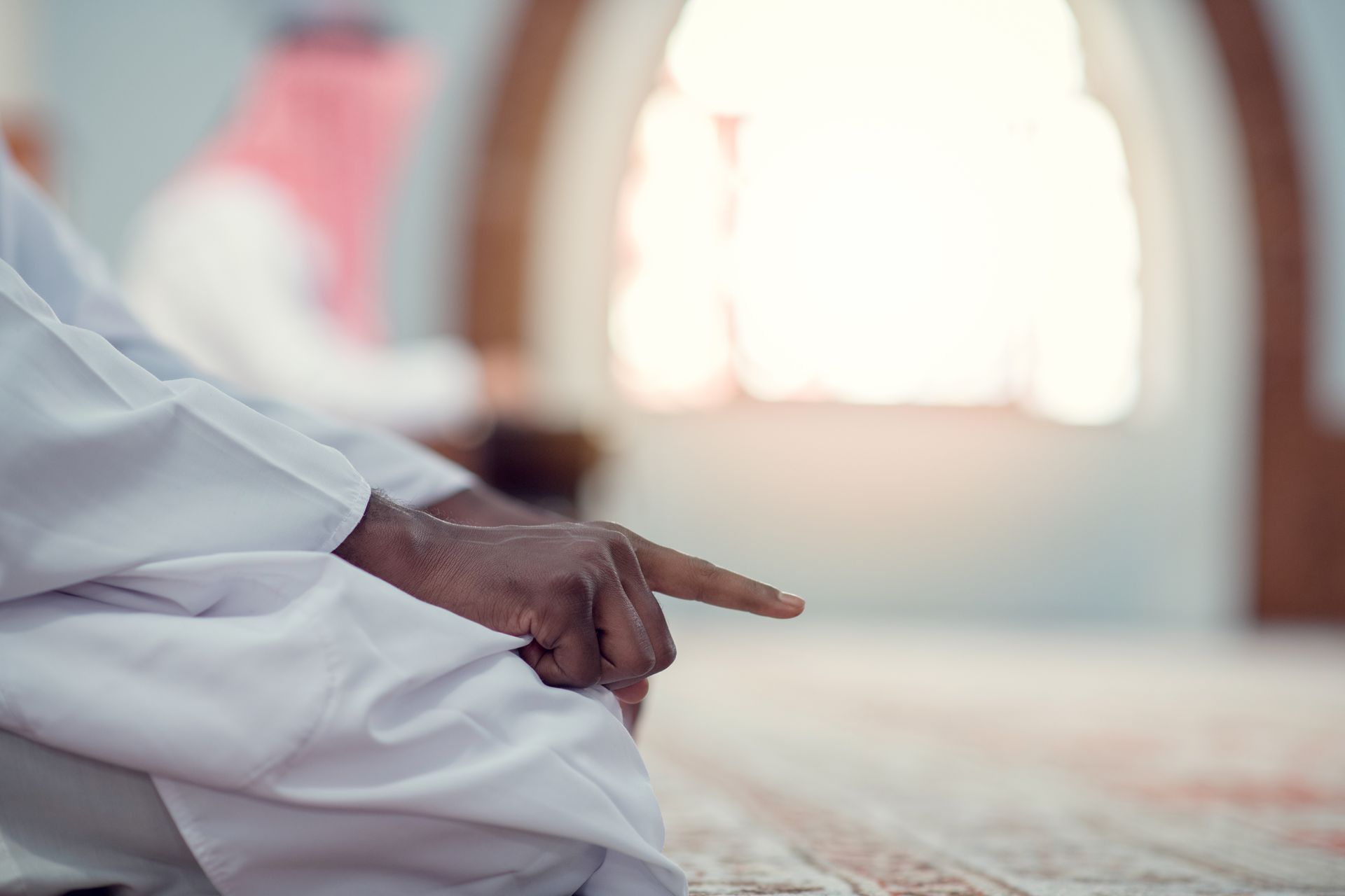 Homme en robe blanche à la peau foncée, la main tendue en signe de prière, à l'intérieur sur un tapis.