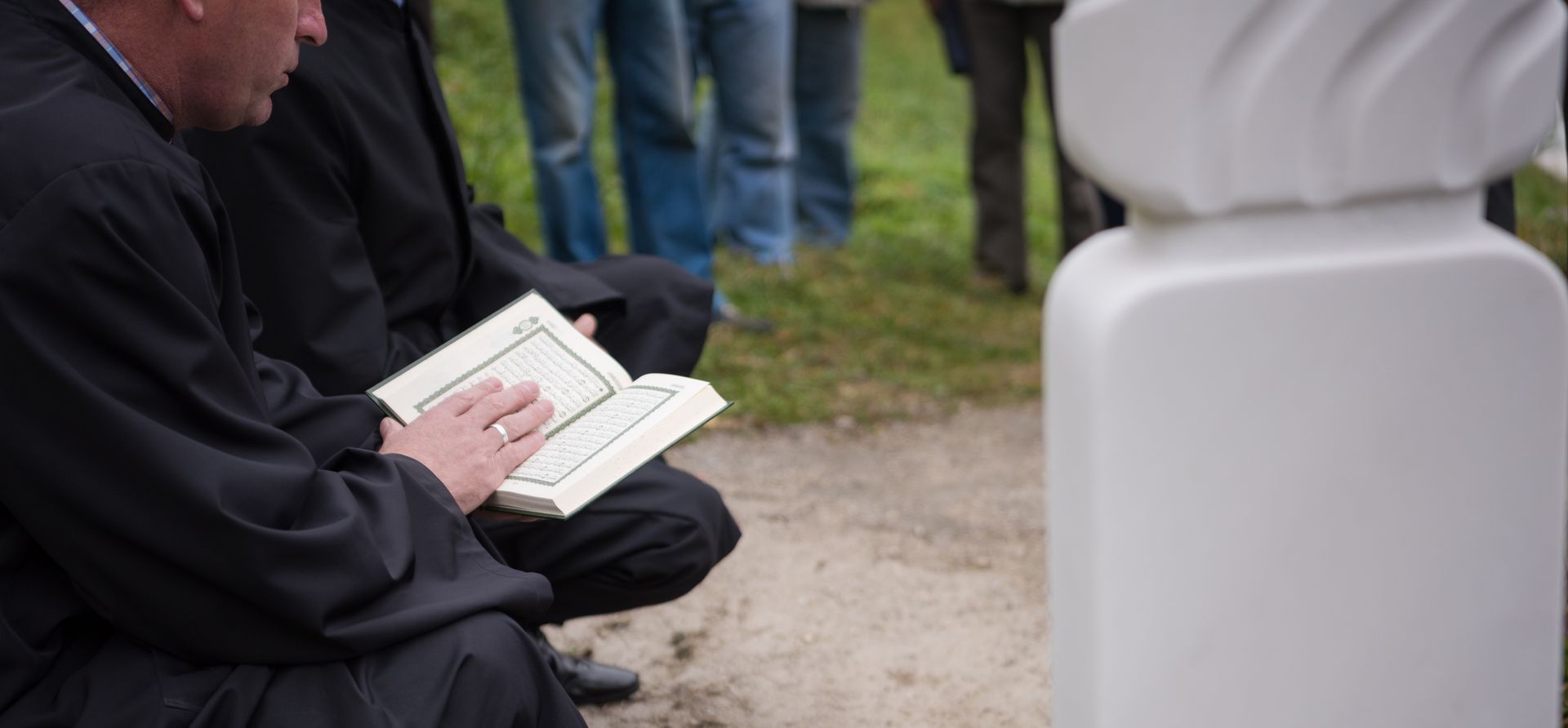 Un homme en noir lisant un livre près d'une pierre tombale blanche avec d'autres personnes en arrière-plan.