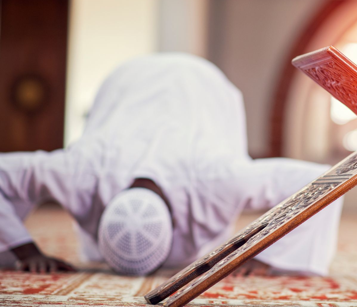 Un homme en vêtements blancs s'incline en prière près d'un présentoir à livres en bois, à l'intérieur d'une mosquée.