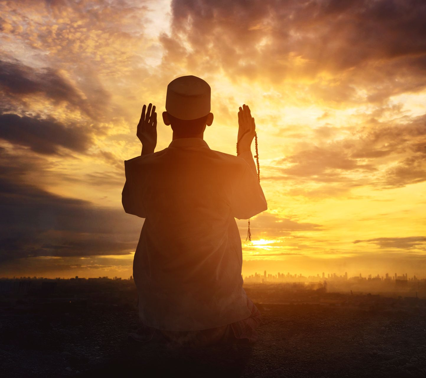 Homme en tenue traditionnelle priant au coucher du soleil, les mains levées.