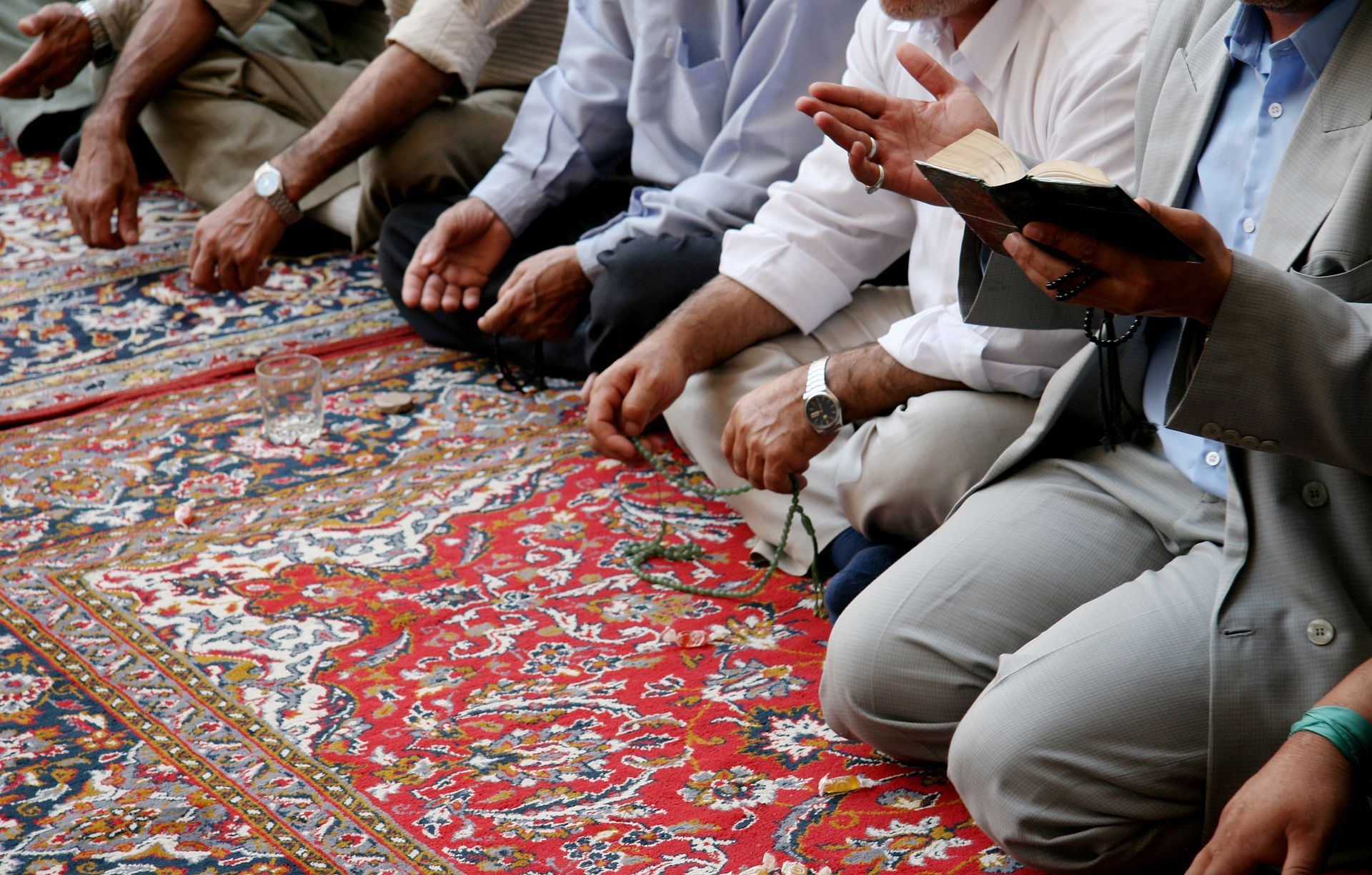 Des hommes en groupe assis sur un tapis à motifs, certains tenant un tasbih, l'un d'eux tenant un livre et levant la main.