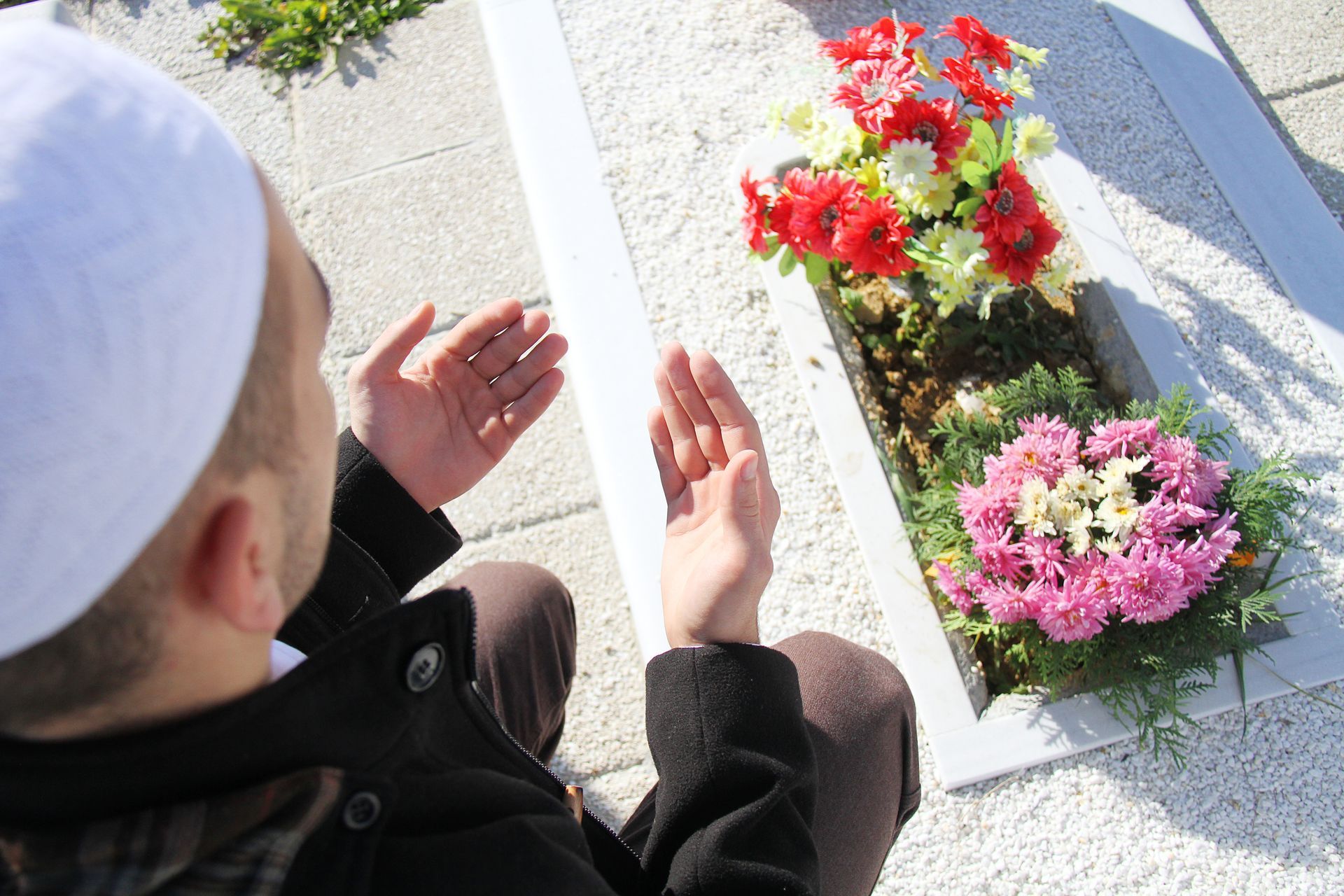 Un homme portant une casquette blanche prie sur une tombe avec des fleurs colorées.