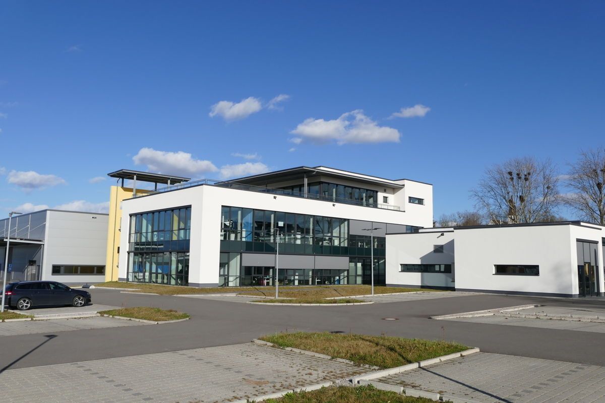 Modernes weißes Bürogebäude mit großen Fenstern und einem kleinen Parkplatz unter strahlend blauem Himmel.