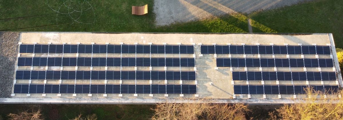 Draufsicht auf ein mit Solarpaneelen bedecktes Gebäudedach, umgeben von Gras und einem kleinen Straßenabschnitt.