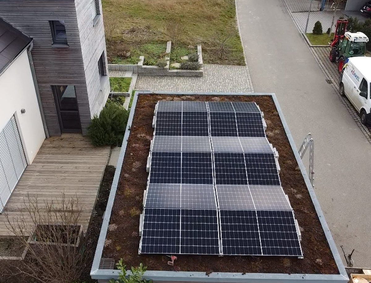 Solarpaneele auf dem Dach eines Gebäudes mit Gründach, neben einer Holzterrasse und einer Straße mit geparkten Fahrzeugen.