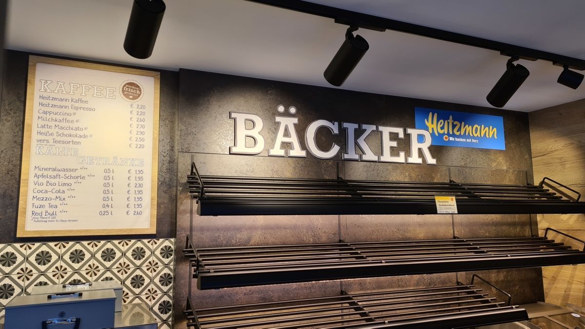 Das Interieur einer Bäckerei mit dem Wort „Bäcker“ über leeren Regalen; an der Wand hängt eine Speisekarte.