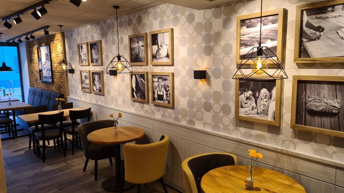 Café-Interieur mit Tischen, Stühlen, gerahmten Fotos an einer gemusterten Wand und Deckenbeleuchtung.