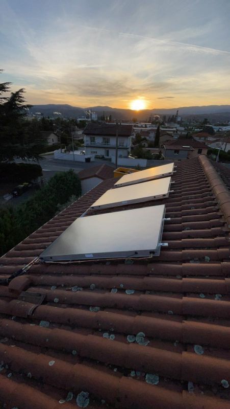 Des panneaux solaires sur un toit de tuiles rouges, avec un coucher de soleil sur une ville en arrière-plan.