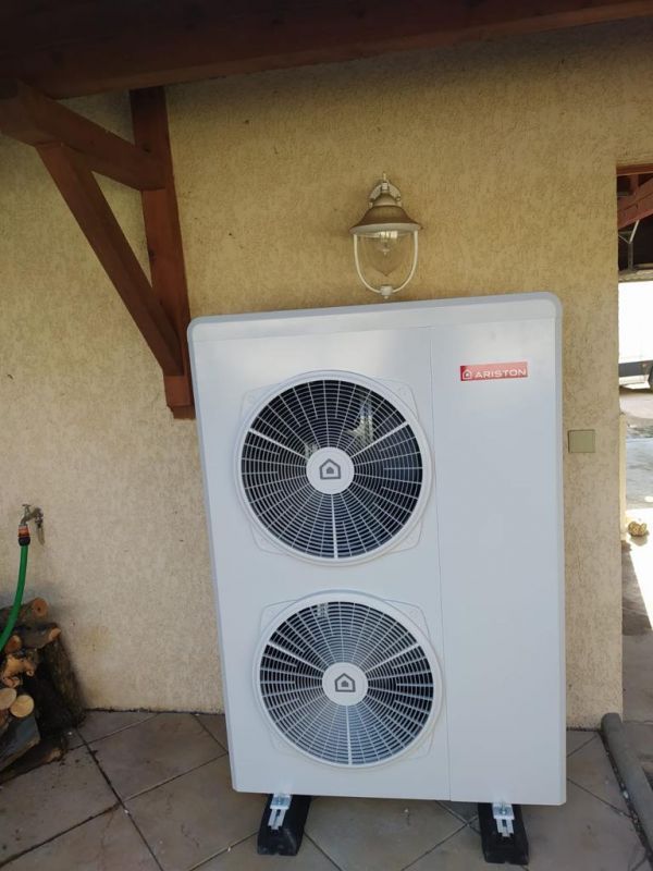 Pompe à chaleur blanche avec deux ventilateurs, montée sur des supports noirs, contre un mur beige, sous un patio couvert.