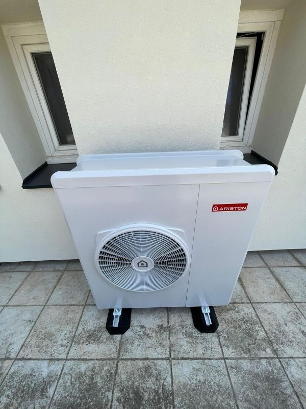 Unité de pompe à chaleur blanche sur une terrasse carrelée, positionnée près d'une fenêtre.