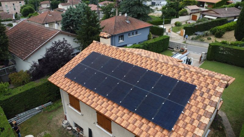 Panneaux solaires sur le toit en tuiles rouges d'une maison à deux étages, entourée d'autres maisons et de verdure.