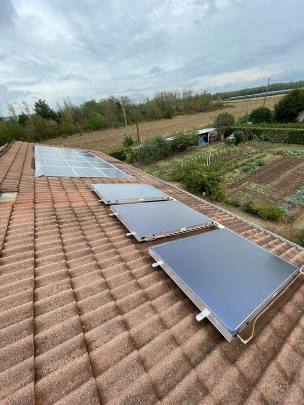 Panneaux solaires et capteurs sur un toit de tuiles en terre cuite. Ciel nuageux, terres agricoles en arrière-plan.
