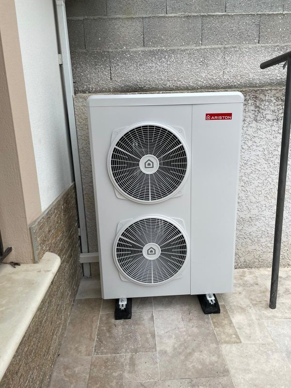 Unité de pompe à chaleur blanche avec deux ventilateurs montés sur des blocs noirs contre un mur gris.