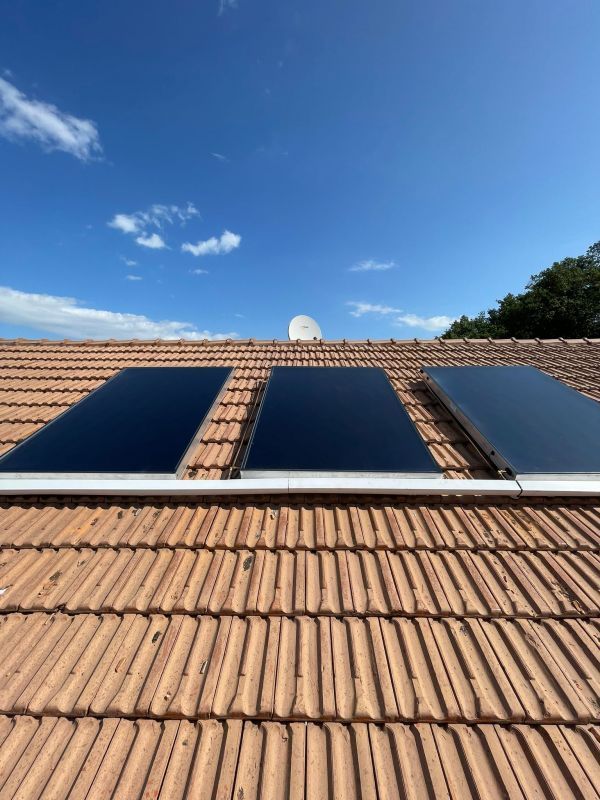 Trois panneaux solaires noirs montés sur un toit de tuiles en terre cuite sous un ciel bleu.