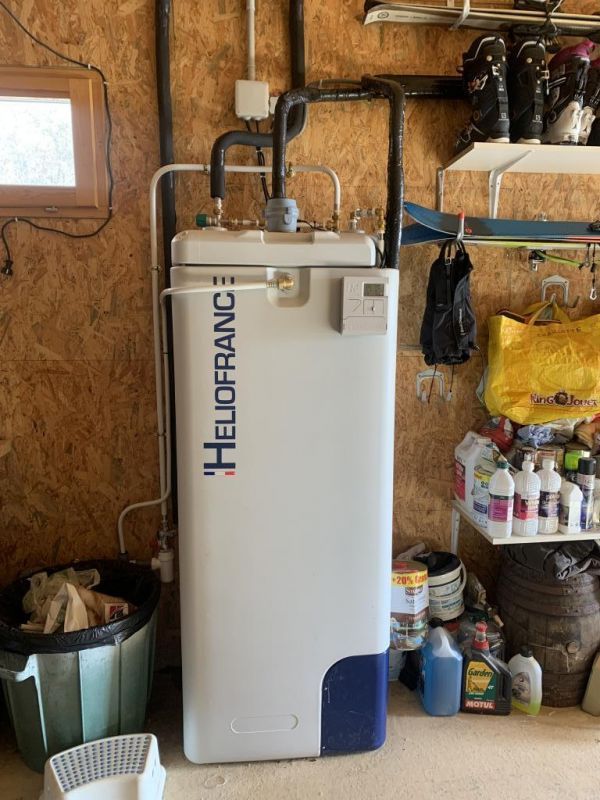 Un chauffe-eau portant l'inscription HELIOFRANCE se trouve dans une buanderie. Des tuyaux noirs y sont raccordés.