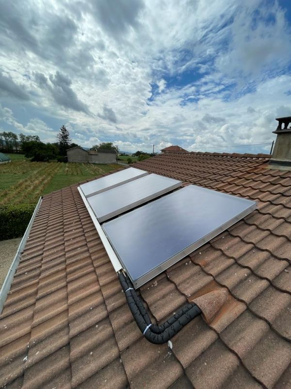 Des panneaux solaires sur un toit de tuiles rouges, avec des tuyaux isolés noirs, se détachant sur un ciel nuageux.