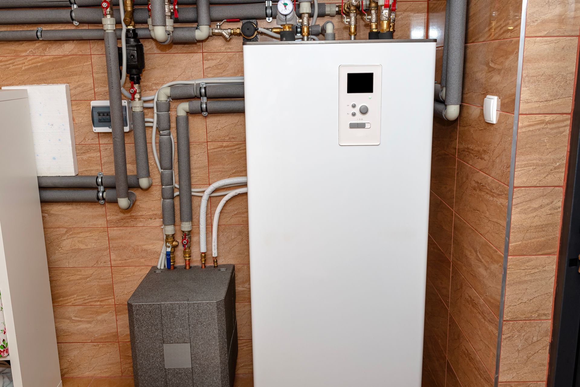 Chauffe-eau blanc et tuyaux isolés gris contre un mur carrelé. Petit boîtier gris sous le chauffe-eau.