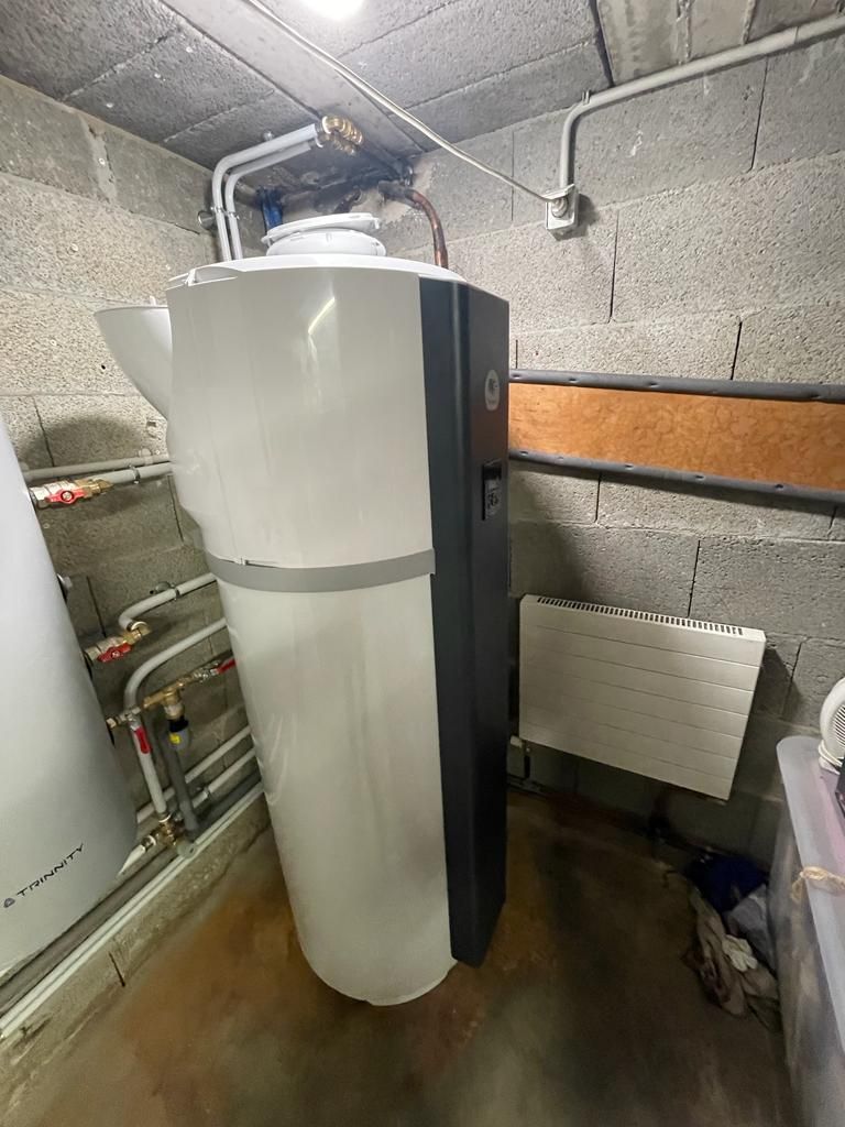 Chauffe-eau cylindrique blanc dans une buanderie aux murs en parpaings et aux tuyaux apparents.