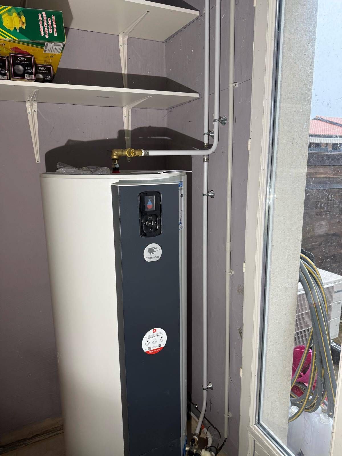 Un grand chauffe-eau blanc à côté d'une fenêtre et des étagères dans une buanderie