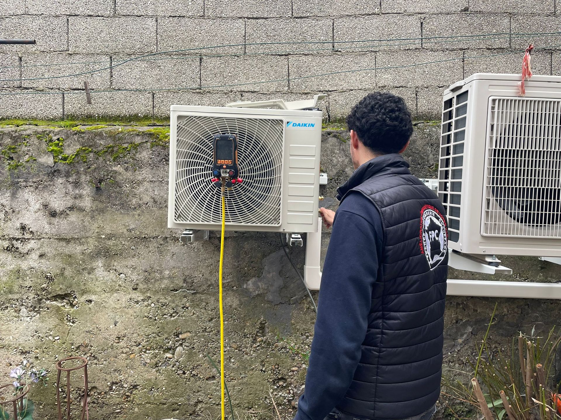 Une personne se tient près d'un boîtier électrique extérieur avec des fils électriques apparents sur un mur en pierre.