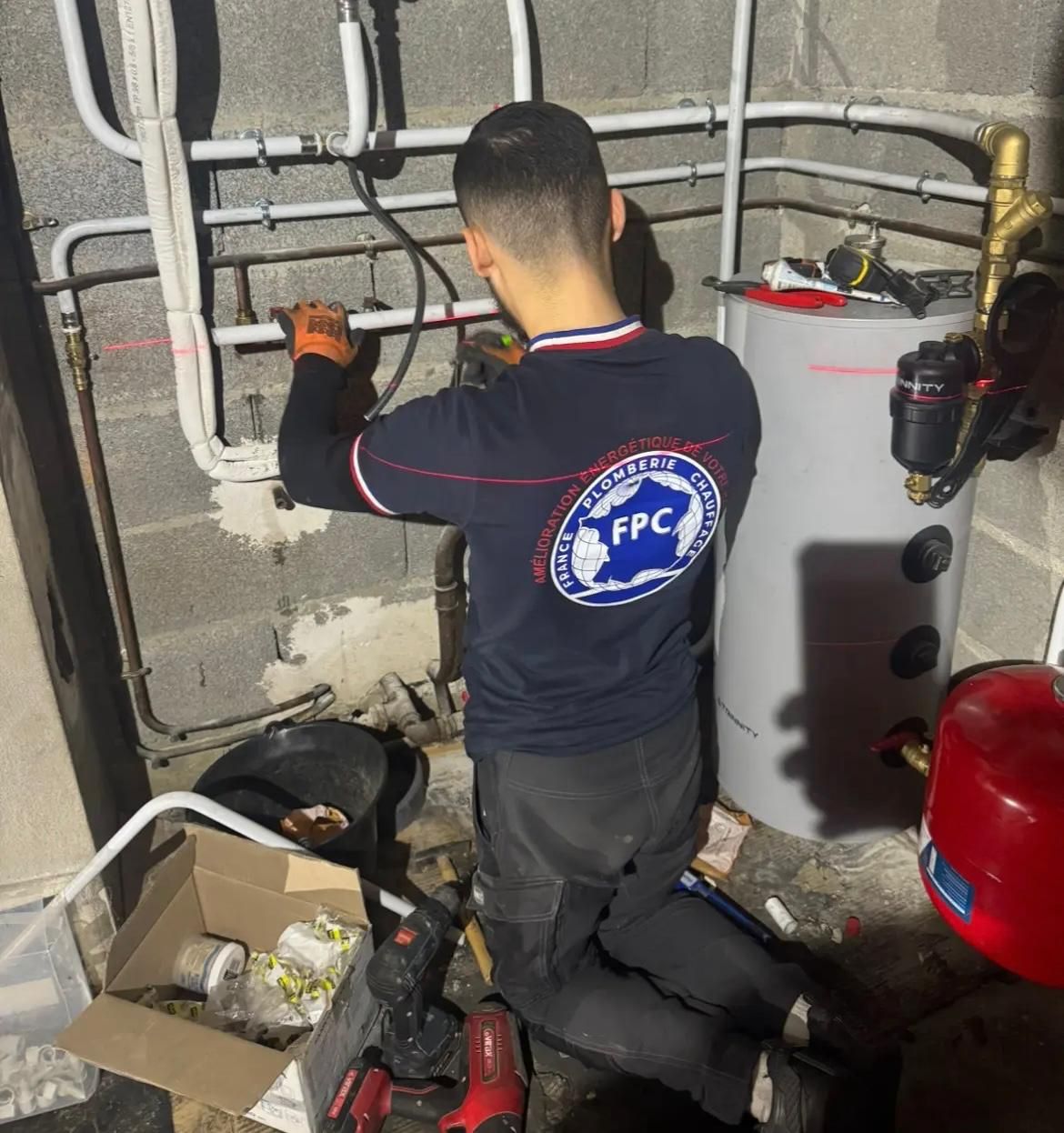 Un technicien répare la plomberie à côté d'un chauffe-eau dans une buanderie, avec des tuyaux et des outils éparpillés à proximité.