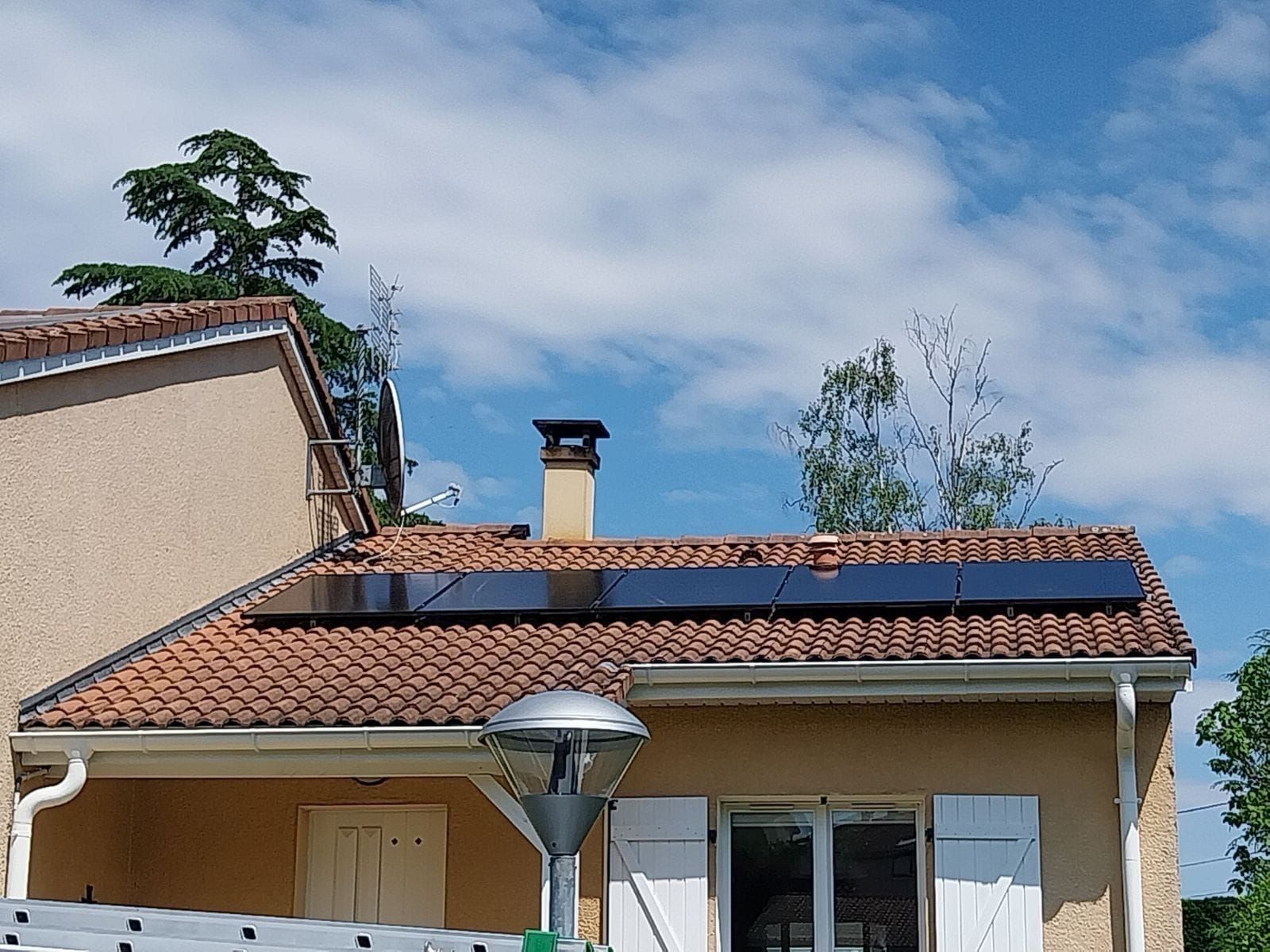 Petite maison au toit de tuiles rouges, panneaux solaires et lampadaire sous un ciel partiellement nuageux