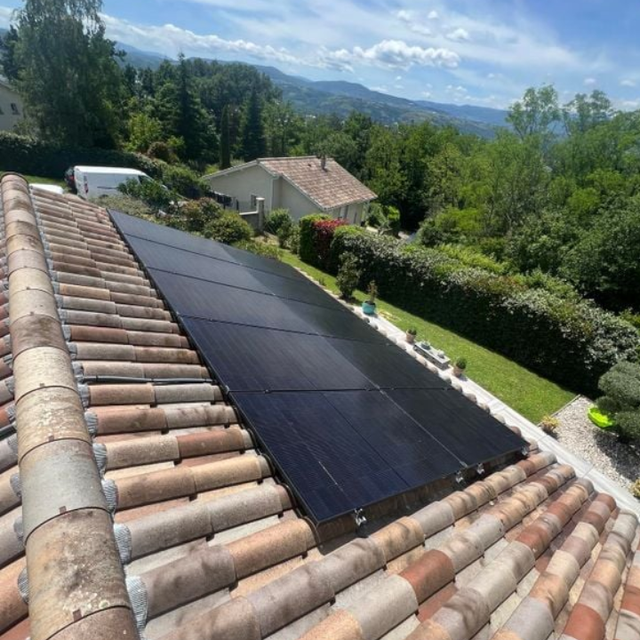 Des panneaux solaires sur un toit de tuiles en terre cuite surplombant une colline verdoyante et des montagnes au loin.