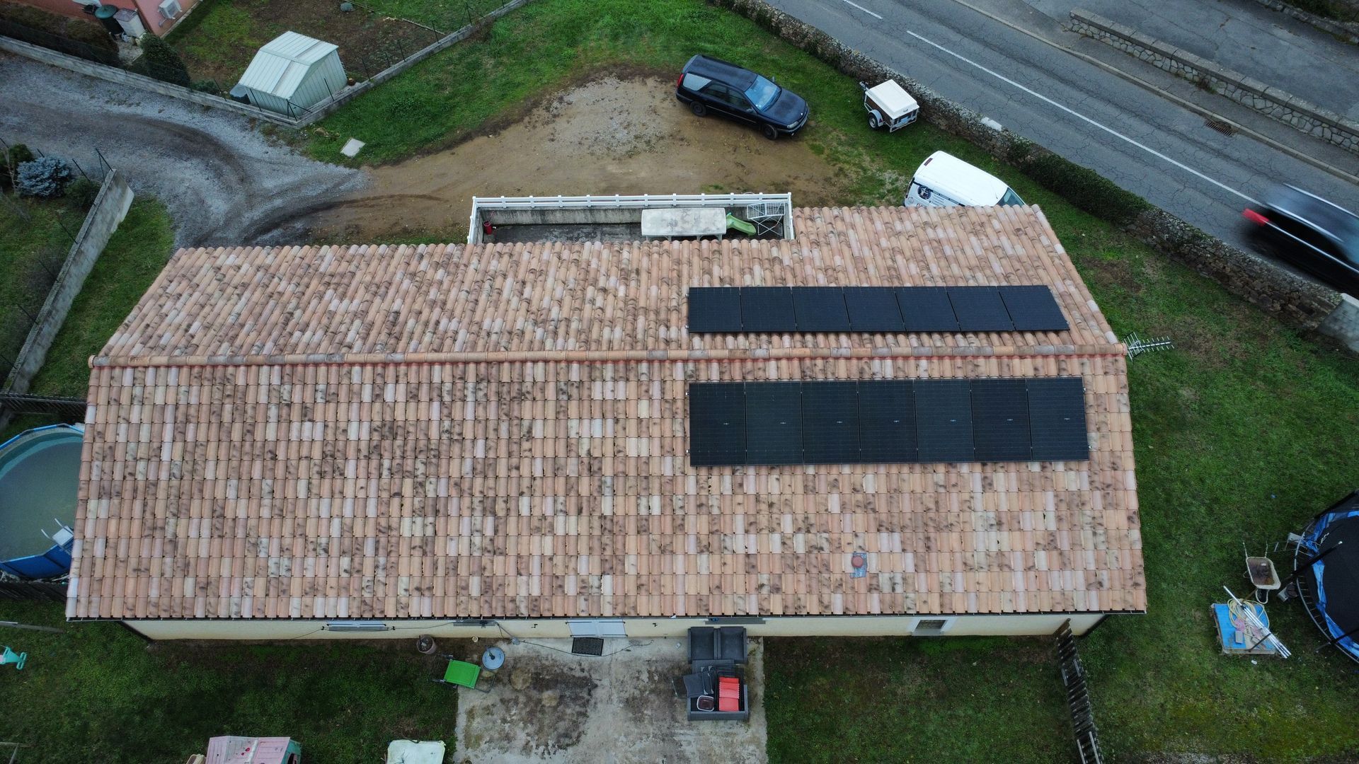 Vue aérienne d'une maison au toit de tuiles équipée de deux panneaux solaires et de voitures garées à proximité.