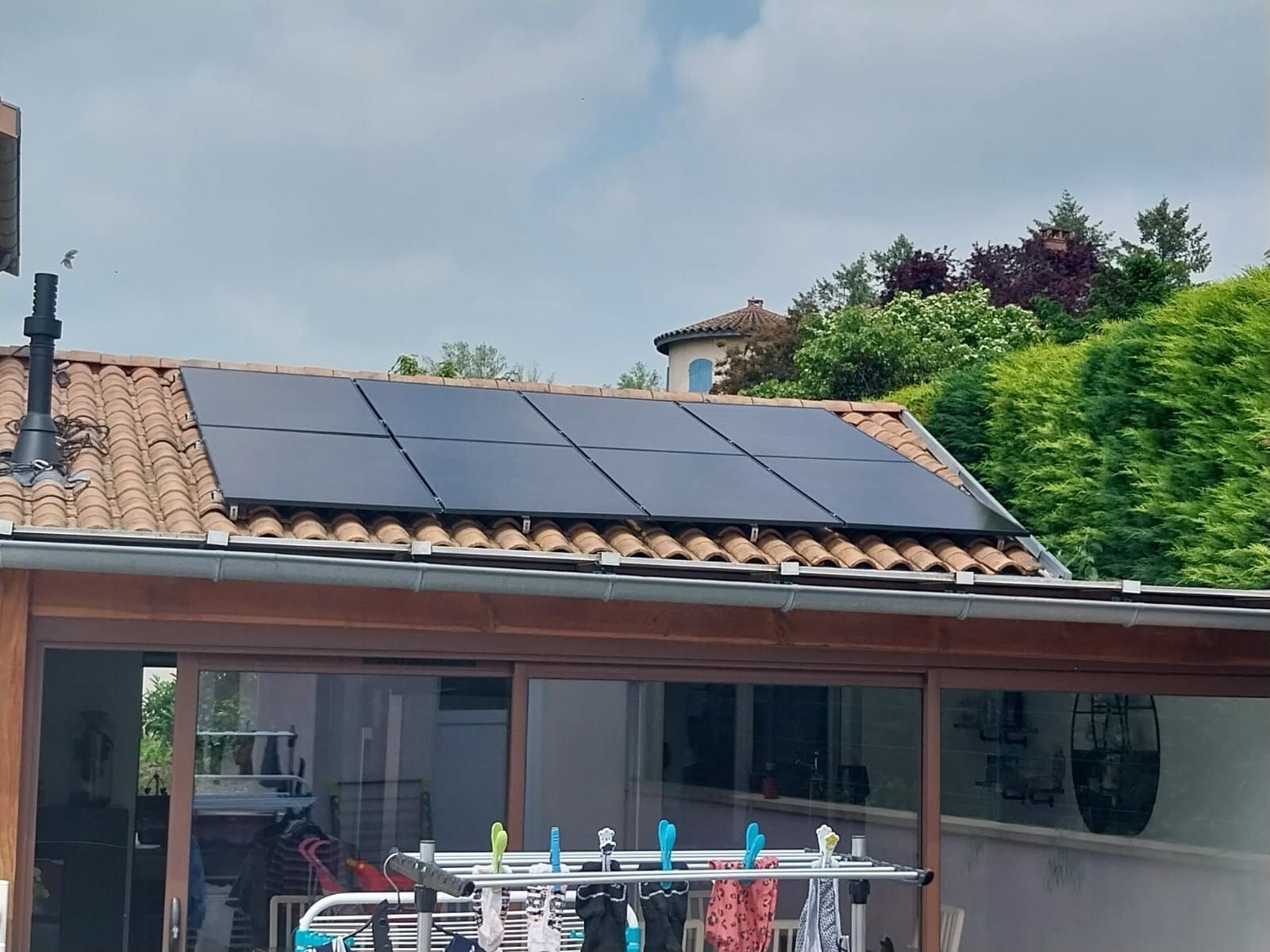 Toit de maison avec panneaux solaires au-dessus d'une terrasse et d'un étendoir à linge, encadré par des arbres et un ciel nuageux