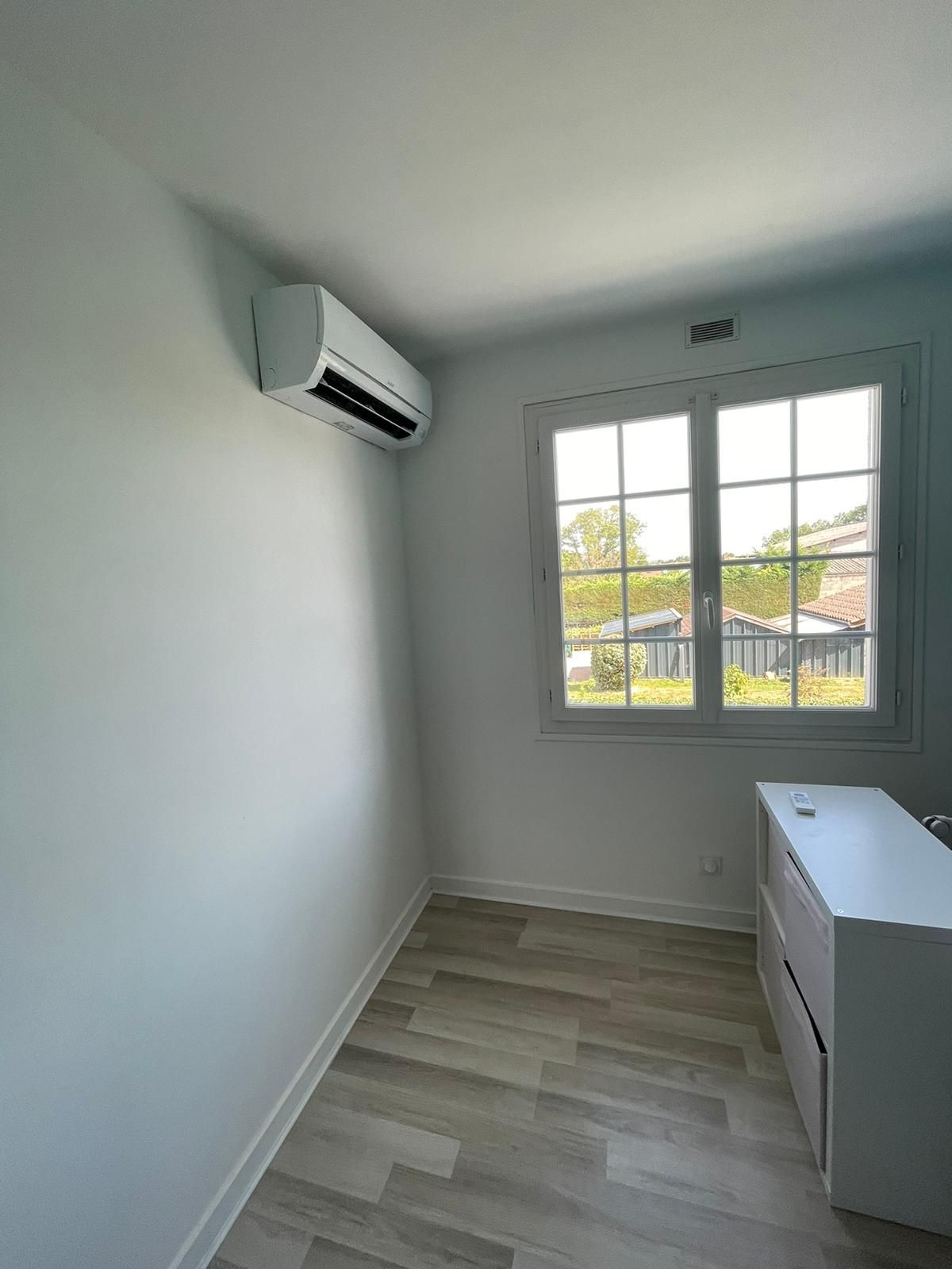 Petite pièce vide aux murs blancs, avec climatiseur mural, fenêtre et armoire blanche.