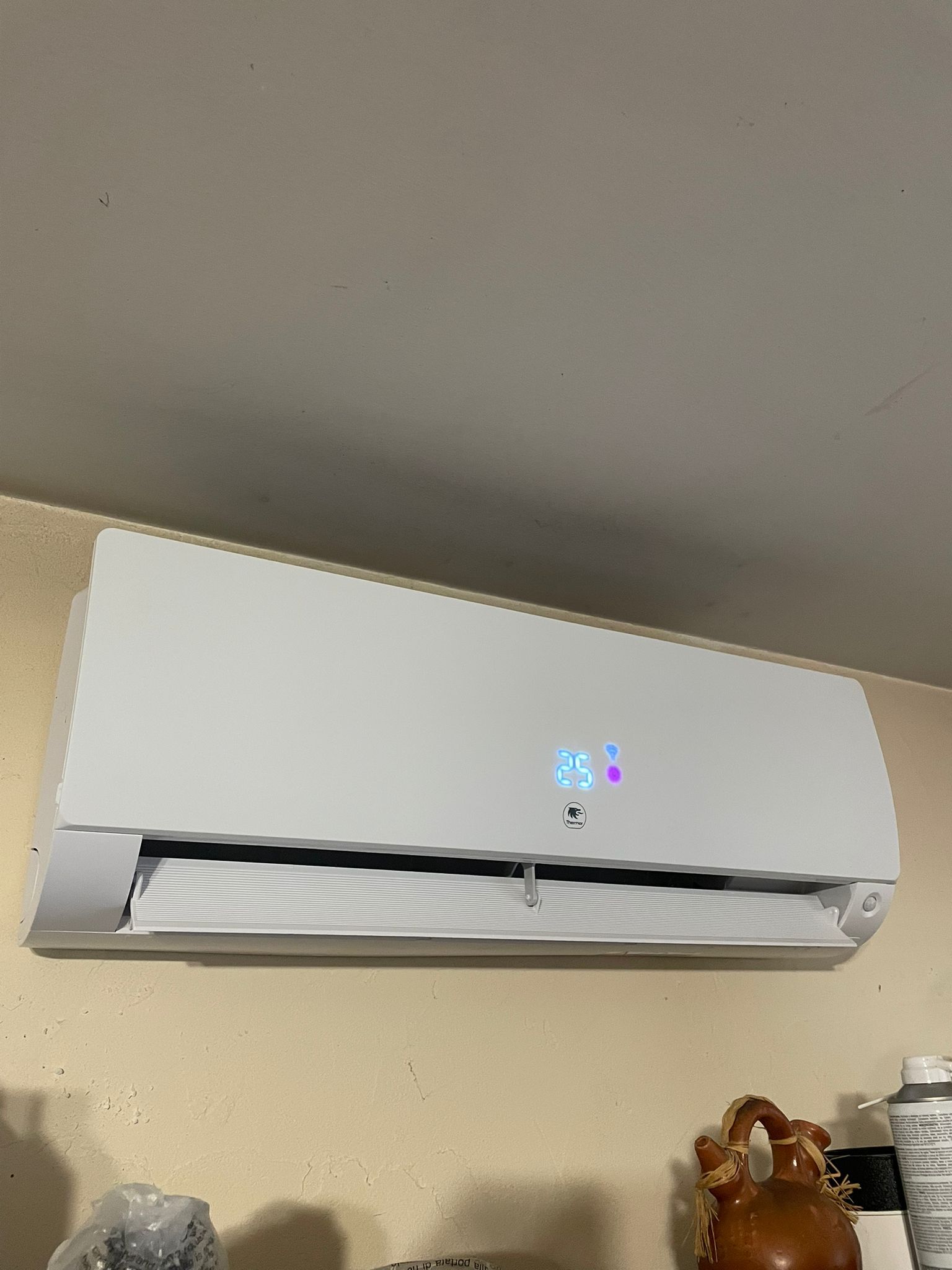 Climatiseur blanc mural au-dessus d'une étagère, avec un petit présentoir éclairé et des figurines en dessous.