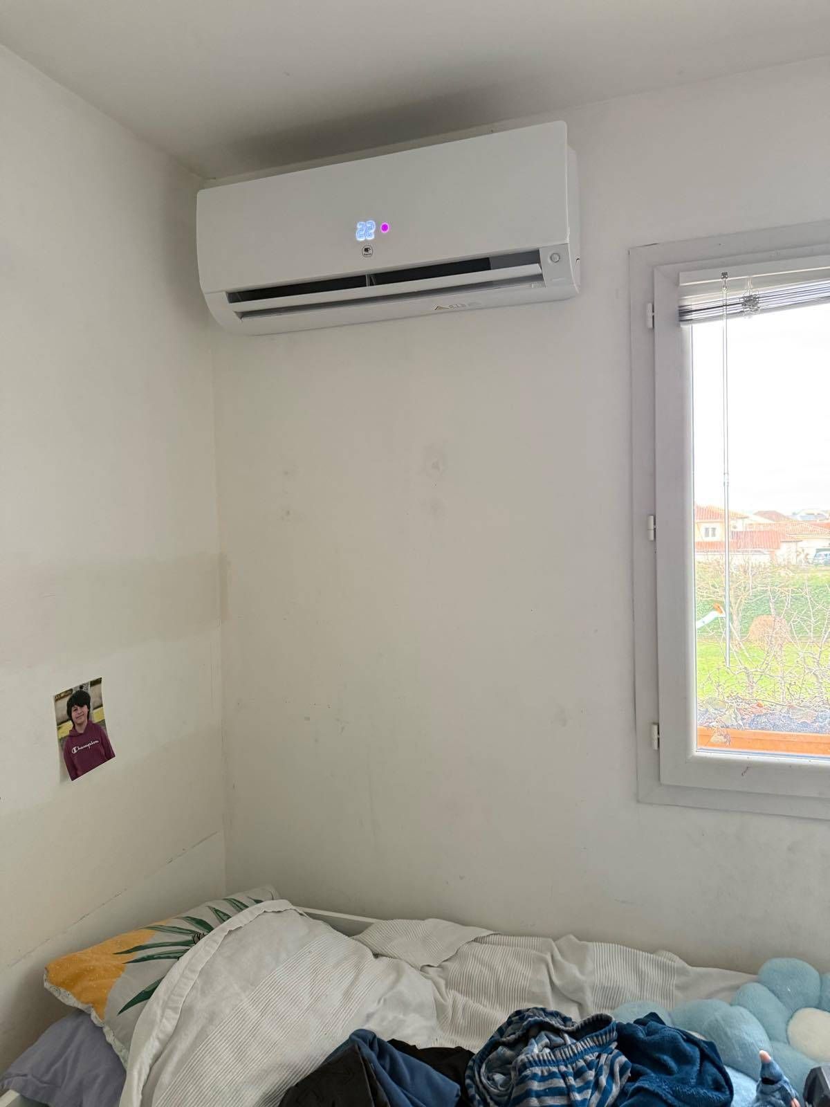 Petite chambre blanche avec climatiseur mural, fenêtre et lit défait avec des vêtements.