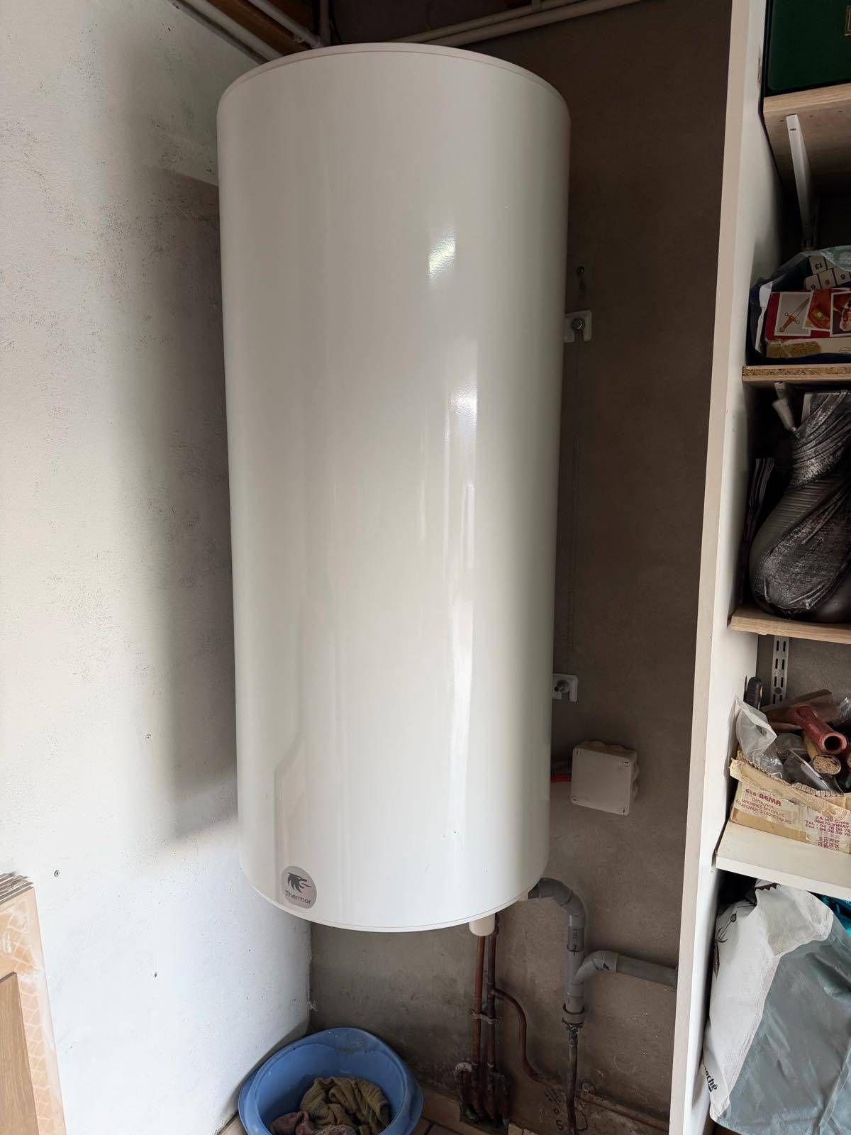 Chauffe-eau cylindrique blanc installé dans un petit placard technique avec des tuyaux et des étagères.