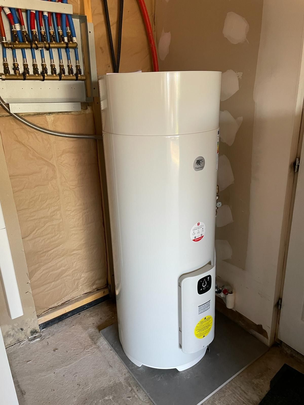 Chauffe-eau blanc installé dans un placard technique, avec tuyaux et câbles apparents.