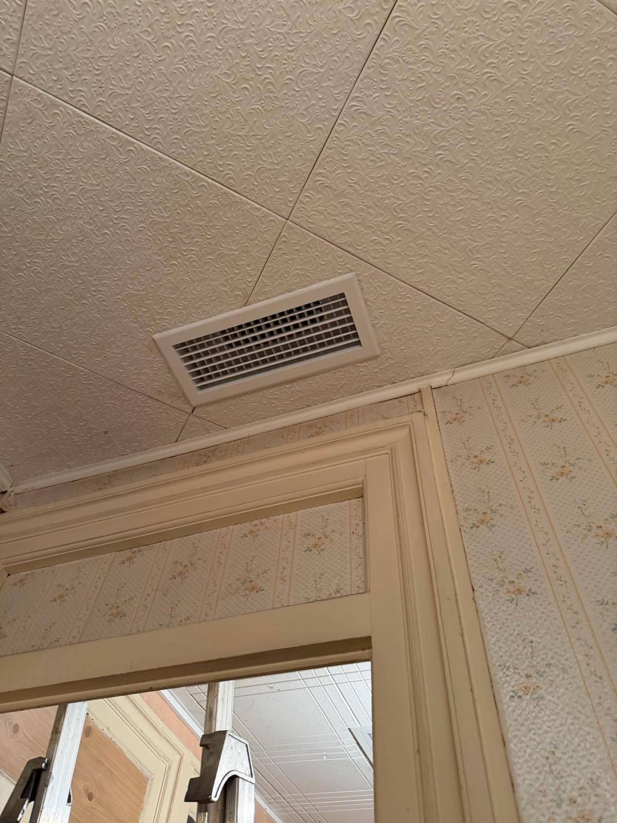 Grille d'aération au plafond au-dessus d'une porte dans une pièce beige avec papier peint à motifs et moulures