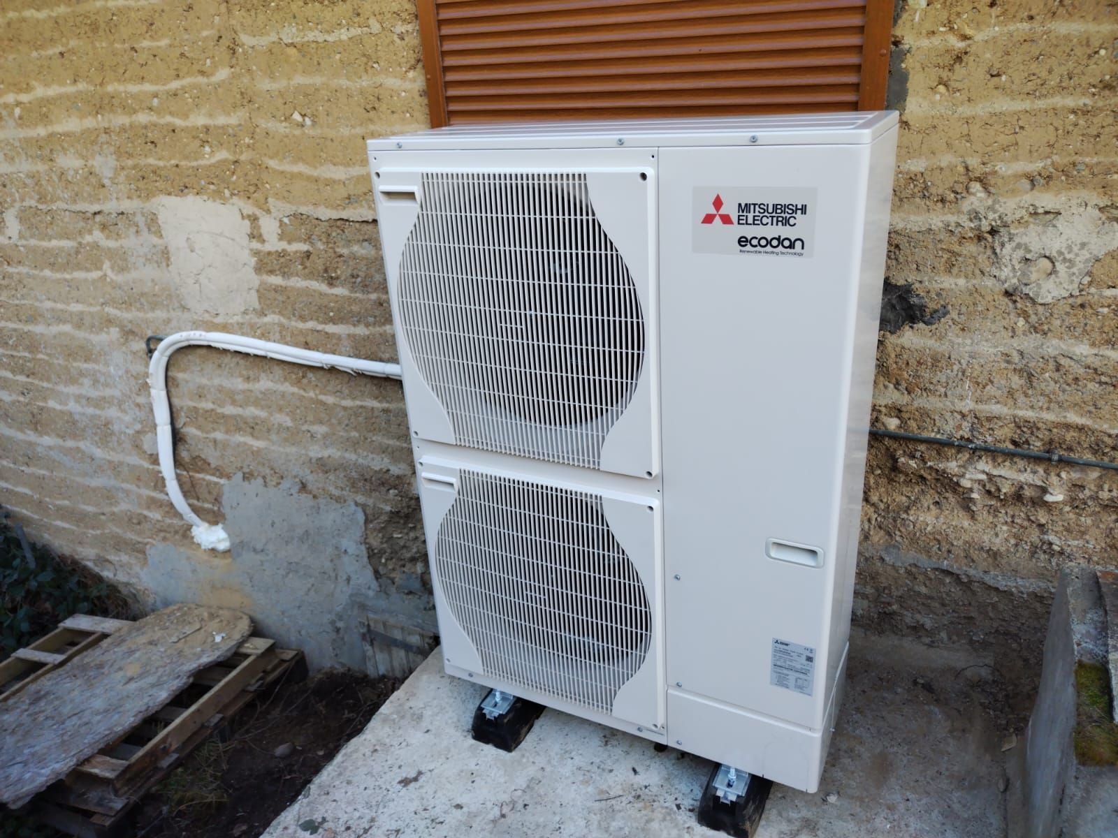 Unité extérieure de climatisation Mitsubishi blanche, fixée contre un mur en pierre, avec tuyauterie apparente.